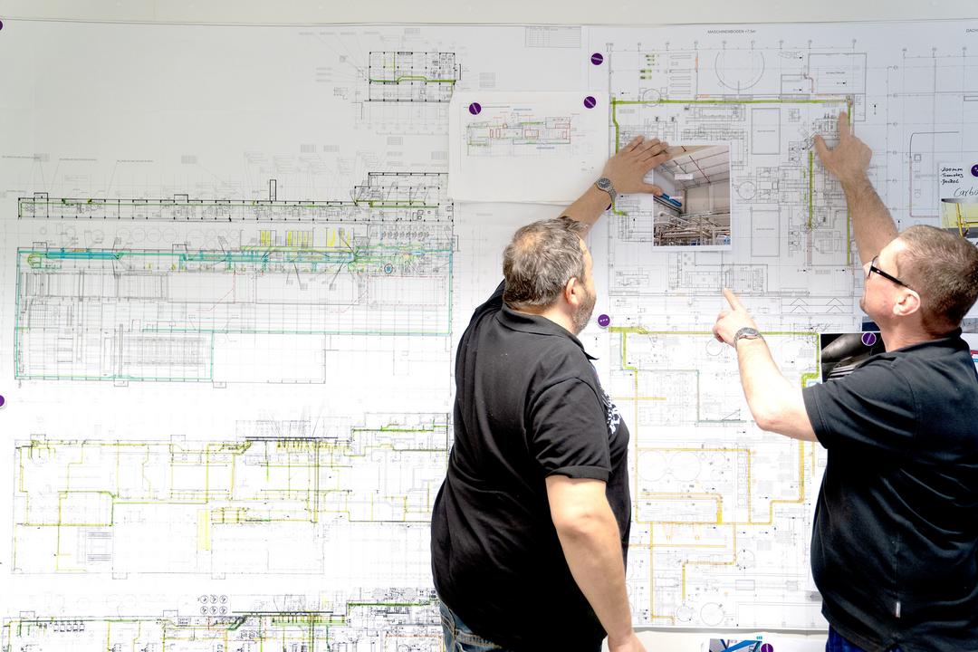 Zwei Männer stehen vor einem großen Plan zur Technischen Gebäudeausrüstung eines Gebäudes