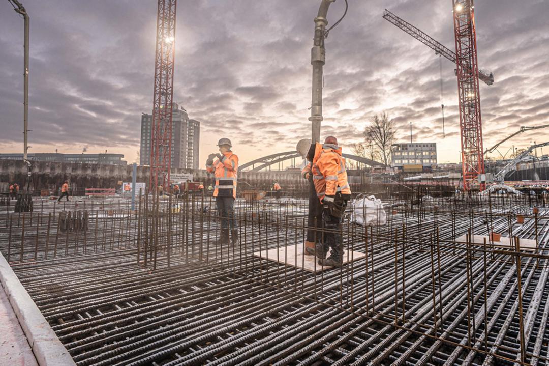 Bauarbeiter auf einer Großbaustelle beim Betonieren einer Bewehrungsplatte bei Sonnenaufgang, umgeben von Kränen und Stahlstäben.